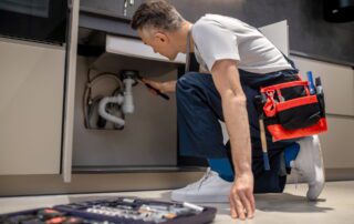 Professional plumber in work uniform kneeling on floor repairing pipes under kitchen sink with toolbox and wrench